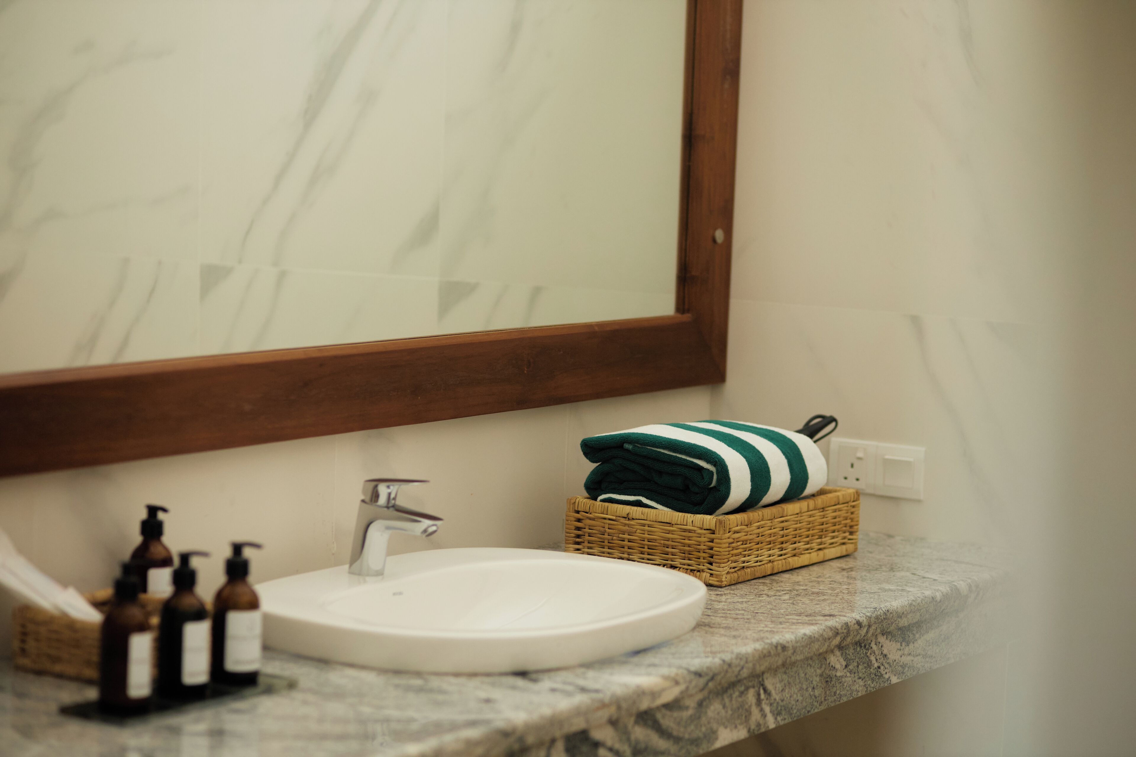 A bathroom with a white sink, a wooden-framed mirror, a wicker basket with a towel, and a marble countertop.