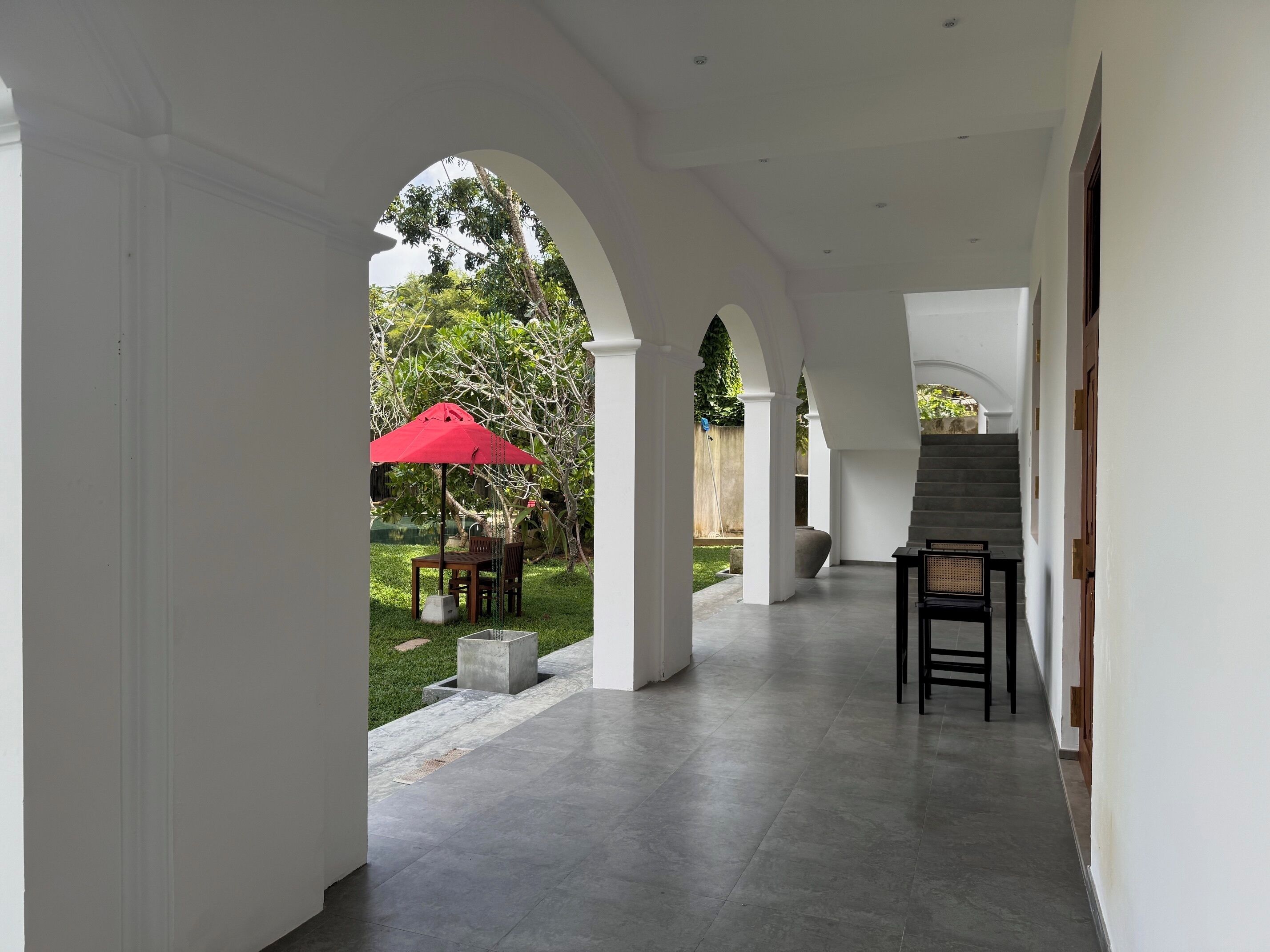 A covered corridor with a view of a garden, a red umbrella, and a staircase.