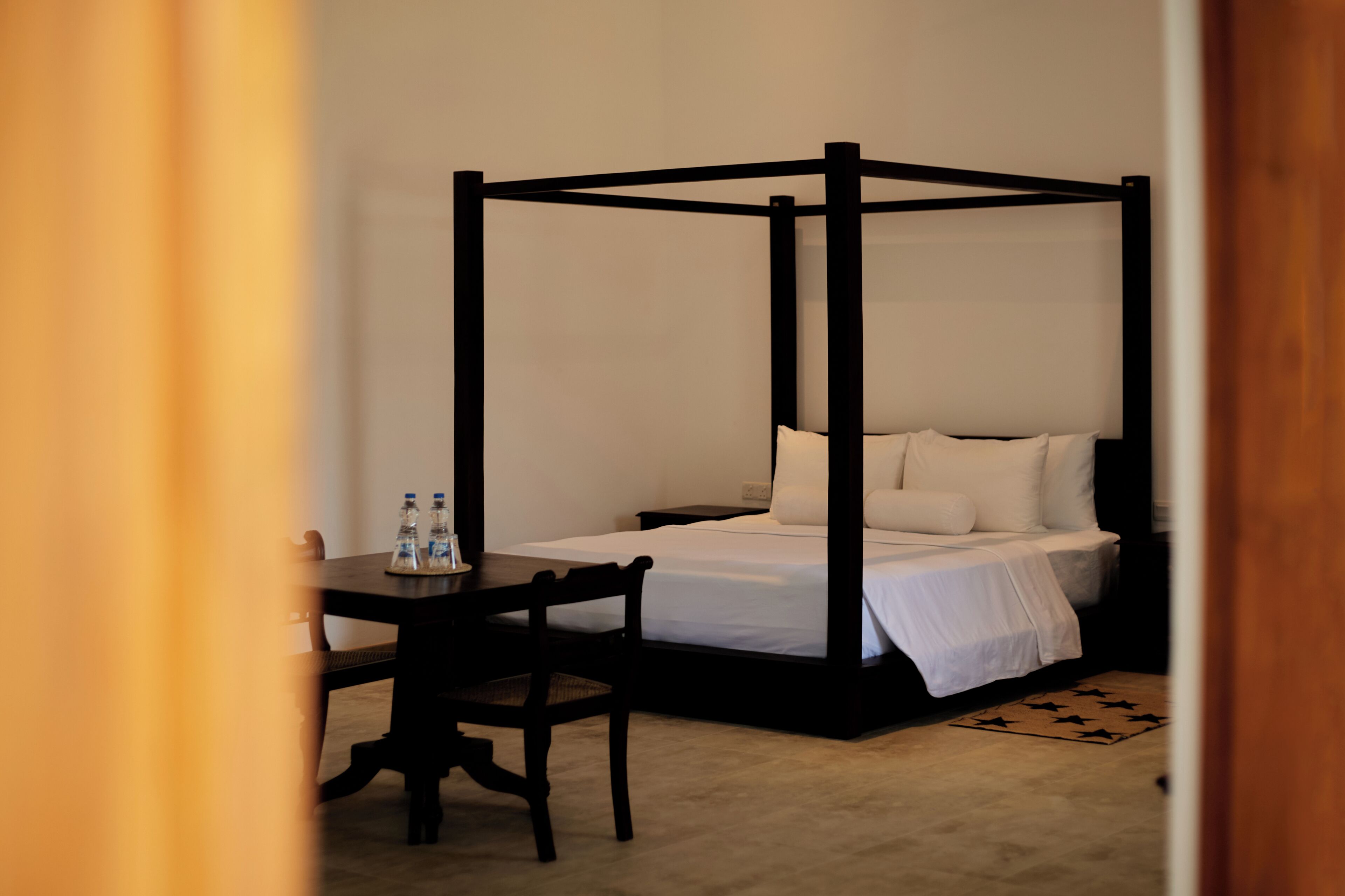 A four-poster bed with white linens, a wooden nightstand, a chair, and two water bottles on the nightstand.
