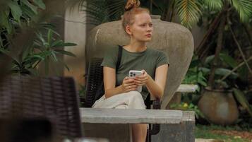 A person seated on a chair in a lush, green outdoor area with various plants and a stone structure.