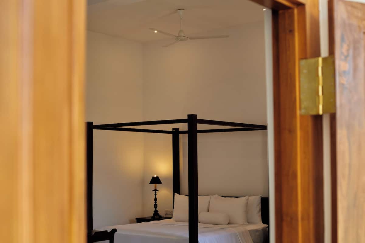 Un letto a baldacchino con lenzuola bianche, un comodino con una lampada e una ventola a soffitto in una stanza con pareti in legno e stipite.