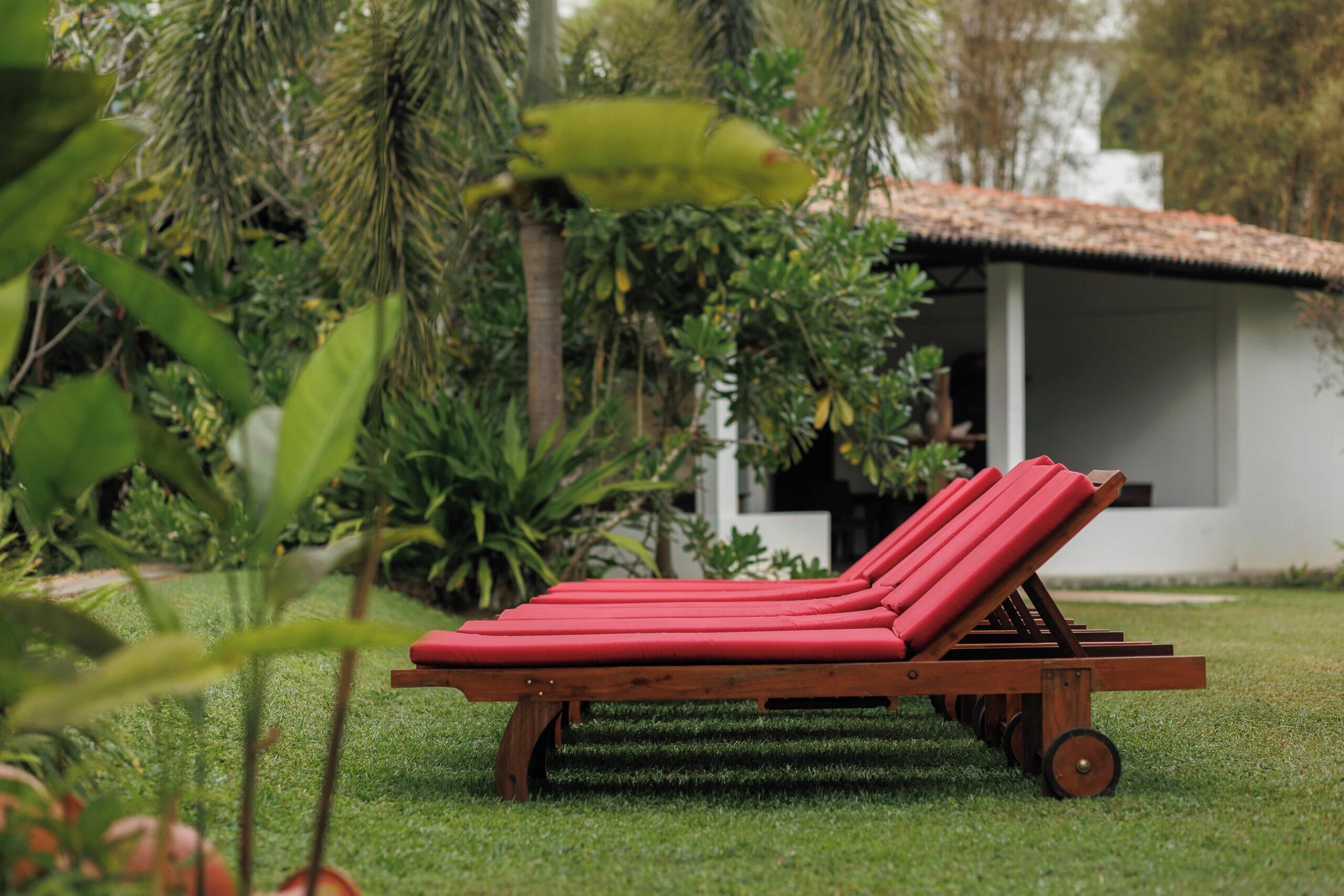 A wooden lounge chair with red cushions in a grassy area with a white building in the background.