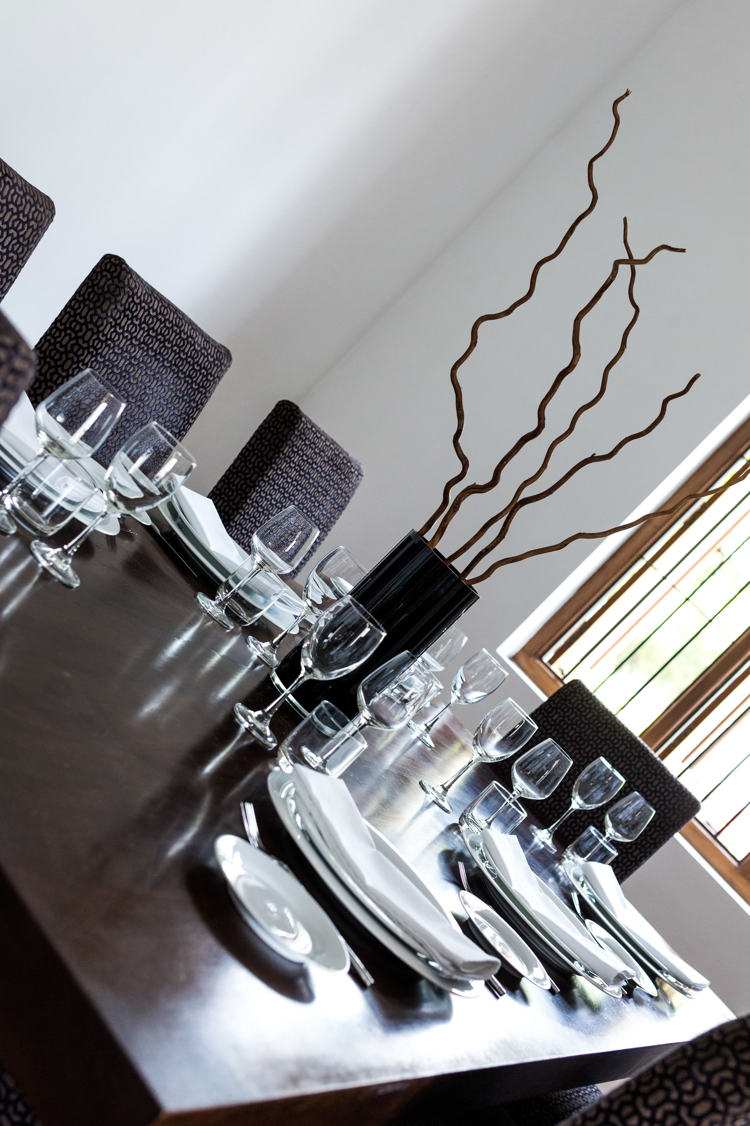 Una mesa de comedor dispuesta con cristalería transparente, platos y cubiertos, con un jarrón de ramas en el centro.