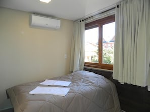 Un dormitorio con cama, unidad de aire acondicionado, ventana con cortinas y vistas a edificios.