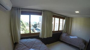 A bedroom with two beds, large windows with curtains, and an air conditioning unit. - Residencial Maria Libardi (Gramado)