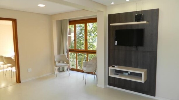 A spacious living room with a large window, a flat-screen TV mounted on a dark wood panel, and two white chairs. - Residencial Maria Libardi (Gramado)