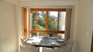 A dining area with a table set for four, large windows with curtains, and a view of greenery outside. - Residencial Maria Libardi (Gramado)