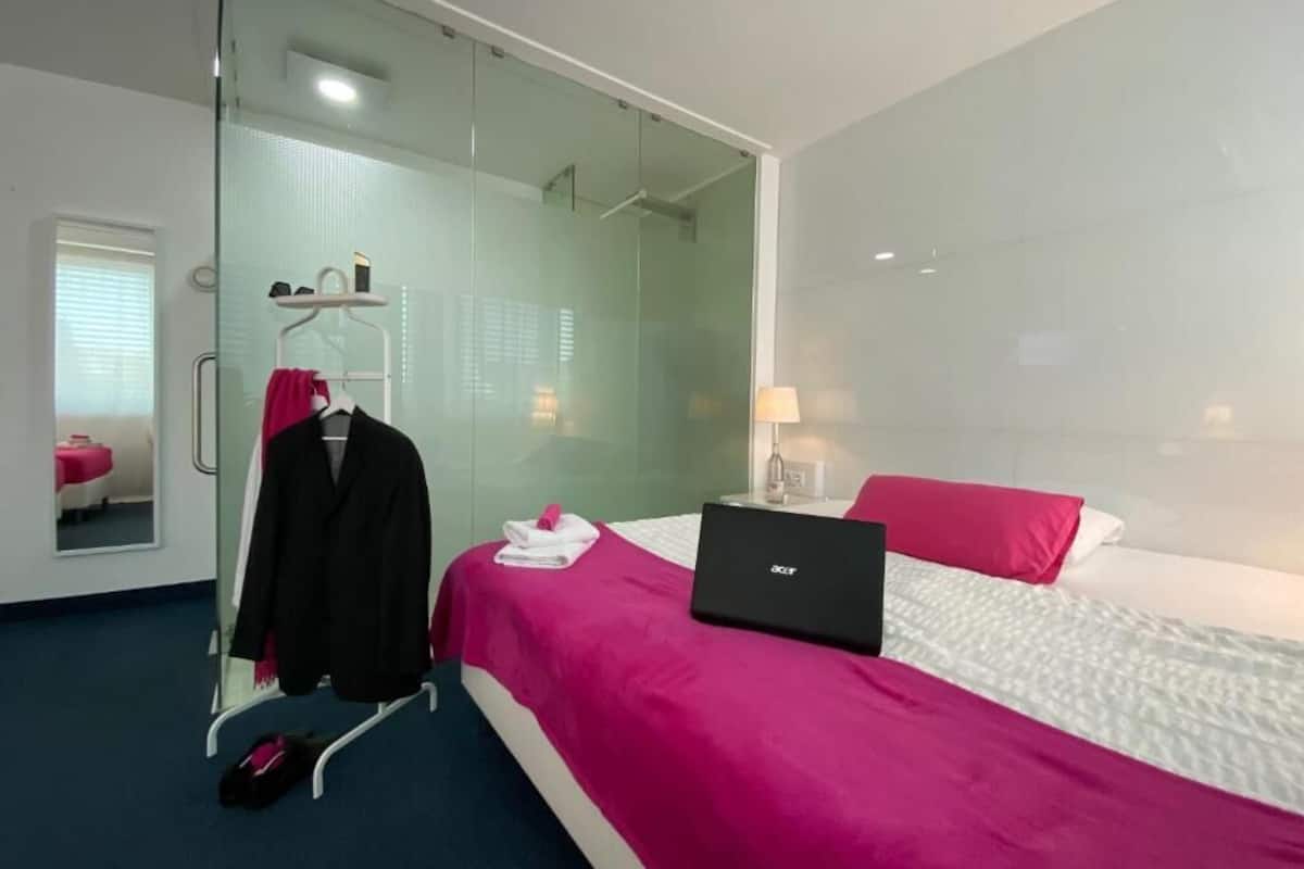 Een moderne hotelkamer met een douchecabine van glas, een bed met een laptop en een roze sprei.