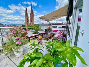 Une terrasse sur le toit offrant une vue sur les deux tours d’une église, aménagée avec des tables et des chaises, et entourée de plantes et de fleurs en pot.