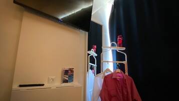 Una habitación con televisión montada en la pared, un estante blanco con control remoto, un perchero con una camisa roja y una ventana con cortina oscura.