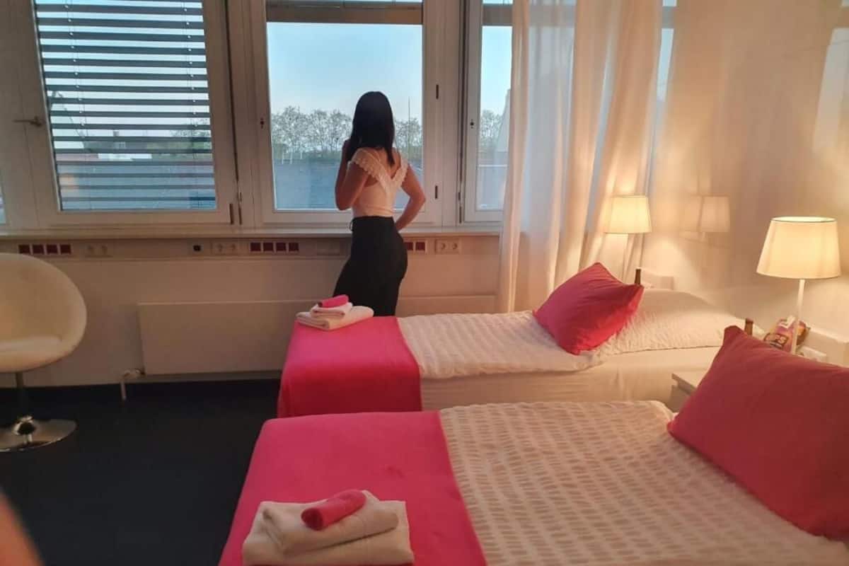 Een hotelkamer met twee bedden, een raam met jaloezieën, een stoel en een lamp.