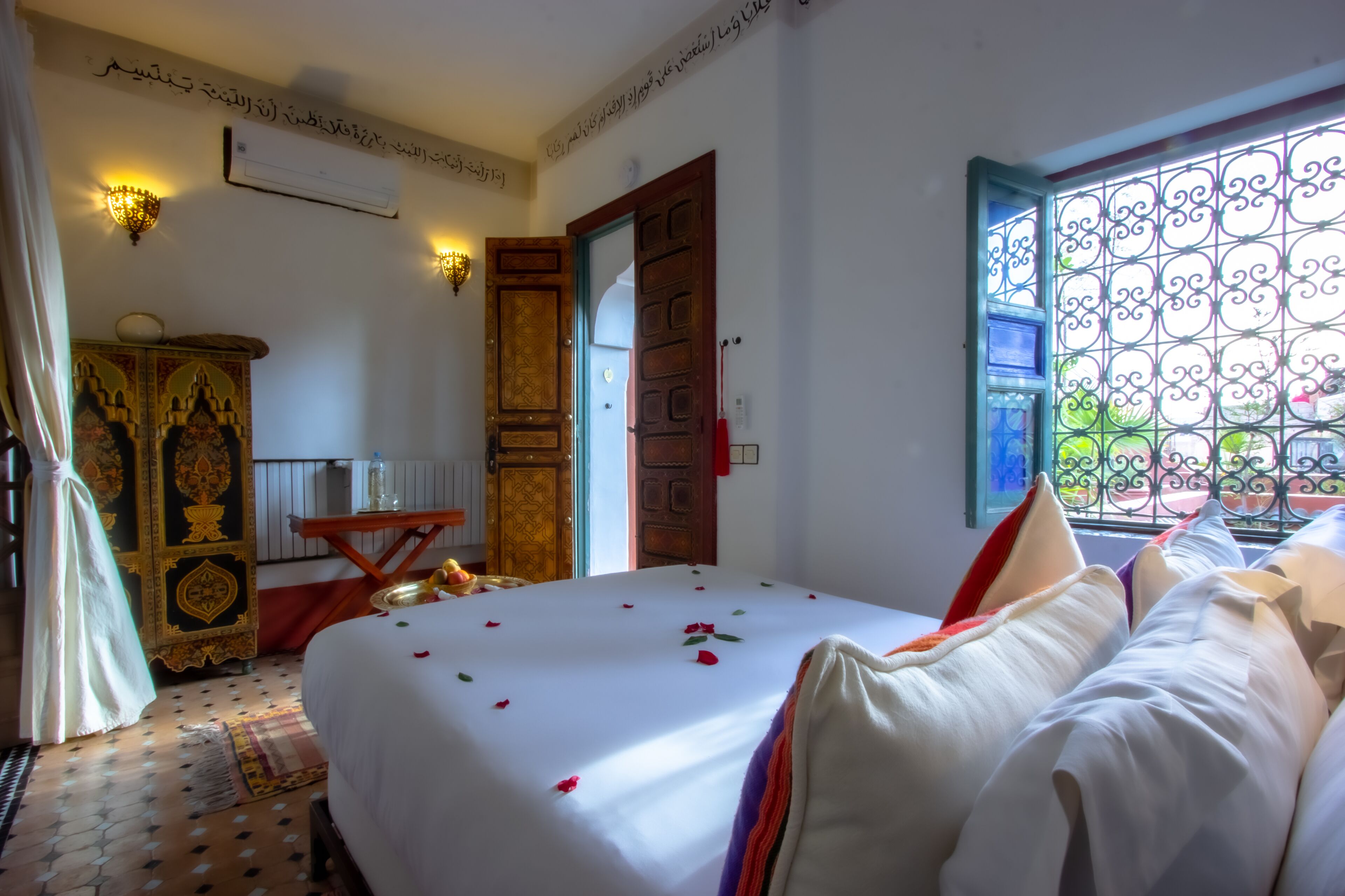 Una habitación de hotel con cama, armario, una mesita y ventana con rejas decorativas de hierro.
