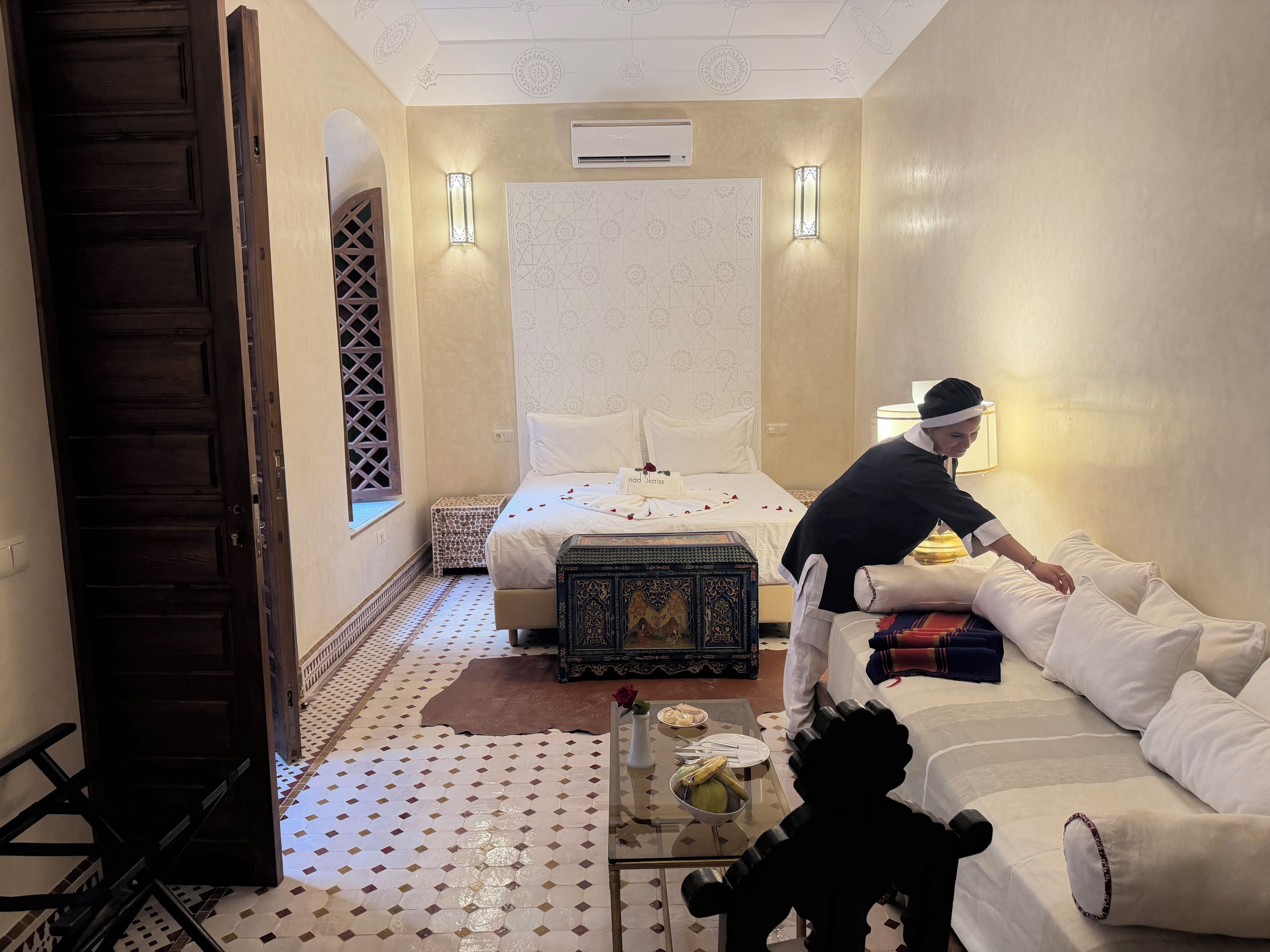 Habitación de hotel con cama, zona de estar con sofá y mesa, almohadas decorativas y aire acondicionado de pared.