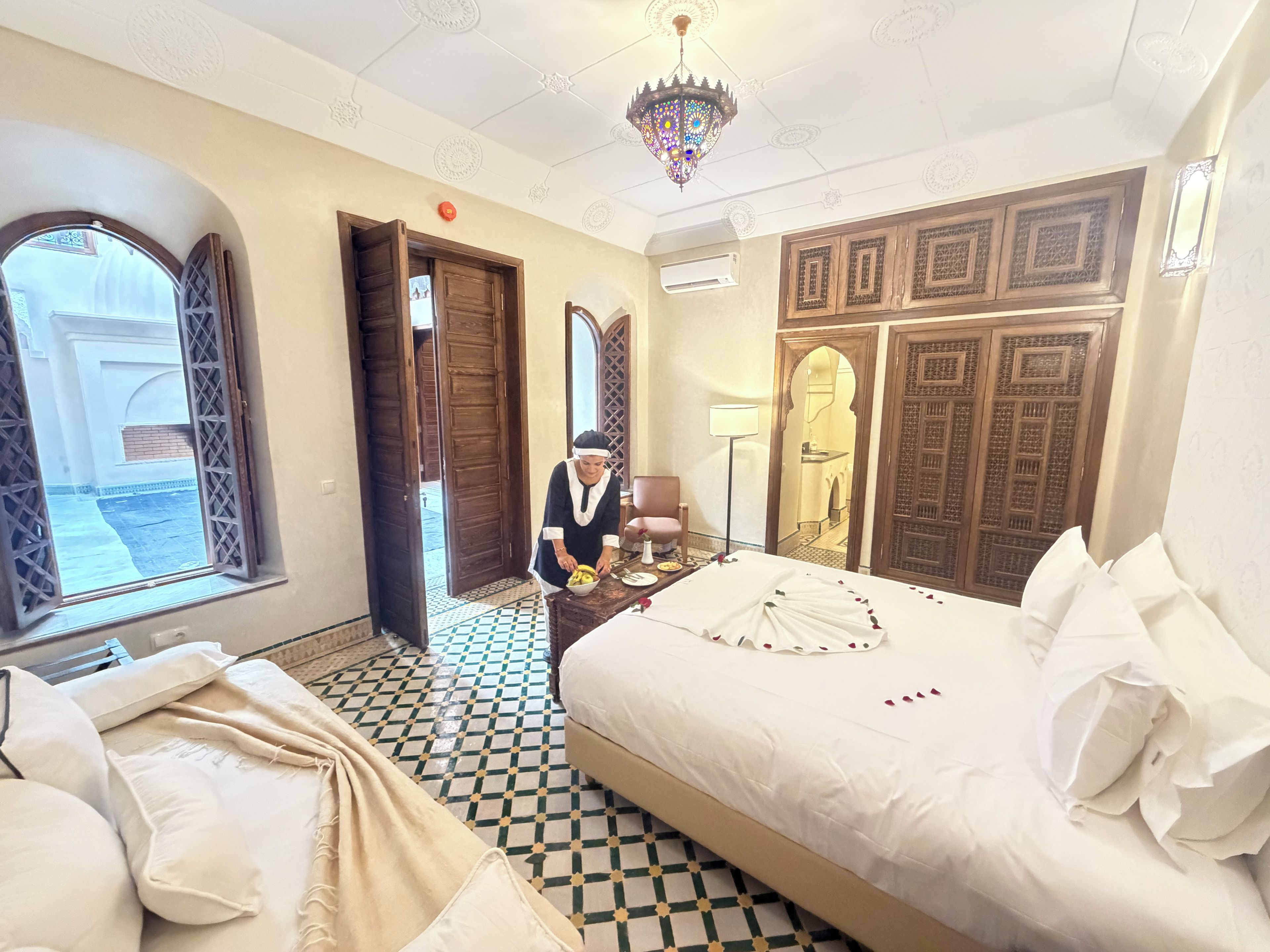 A hotel room with a bed, a chair, a table with a vase, and a chandelier.