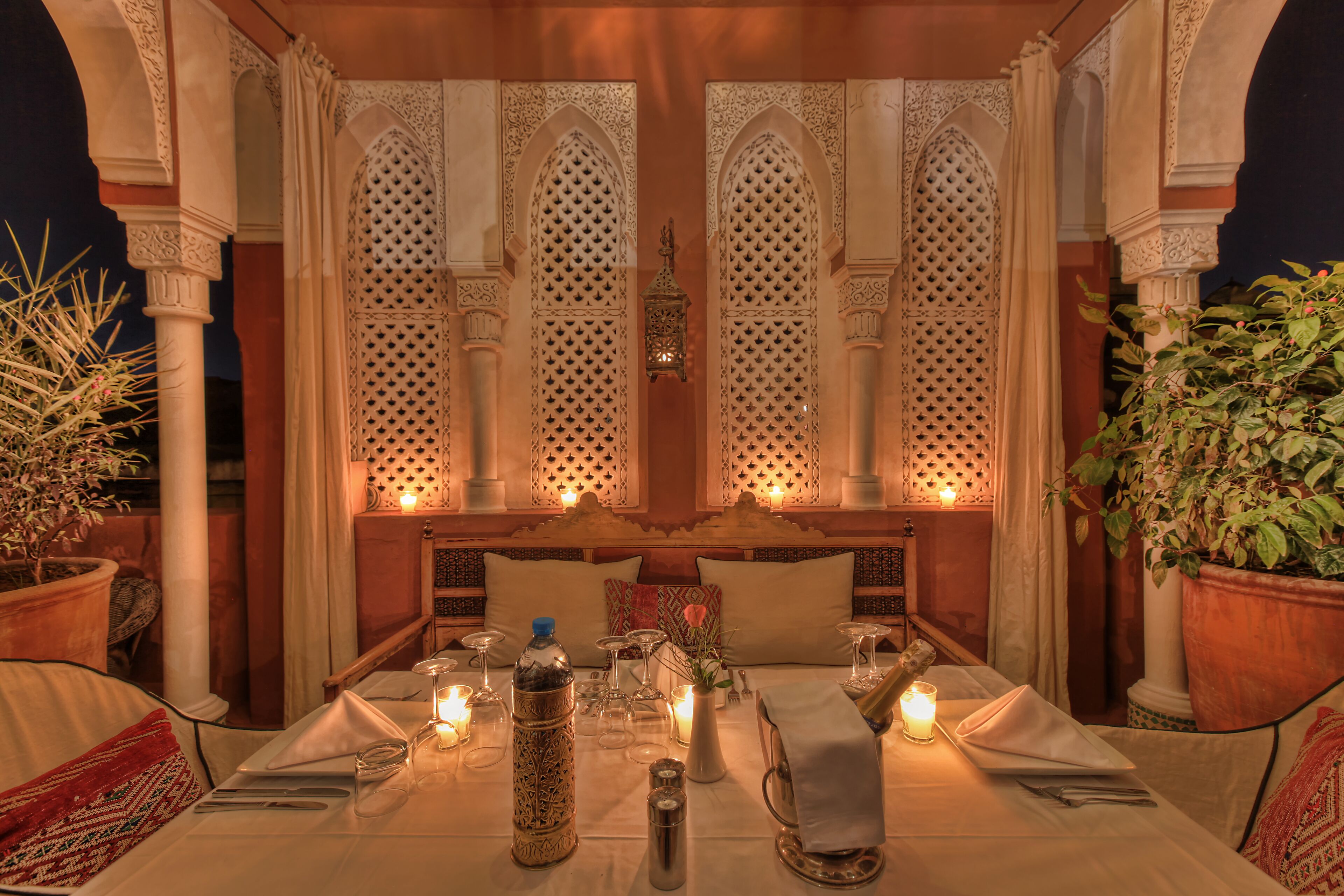 A dining area with a table set for two, featuring a candle-lit ambiance, ornate architectural details, and potted plants.