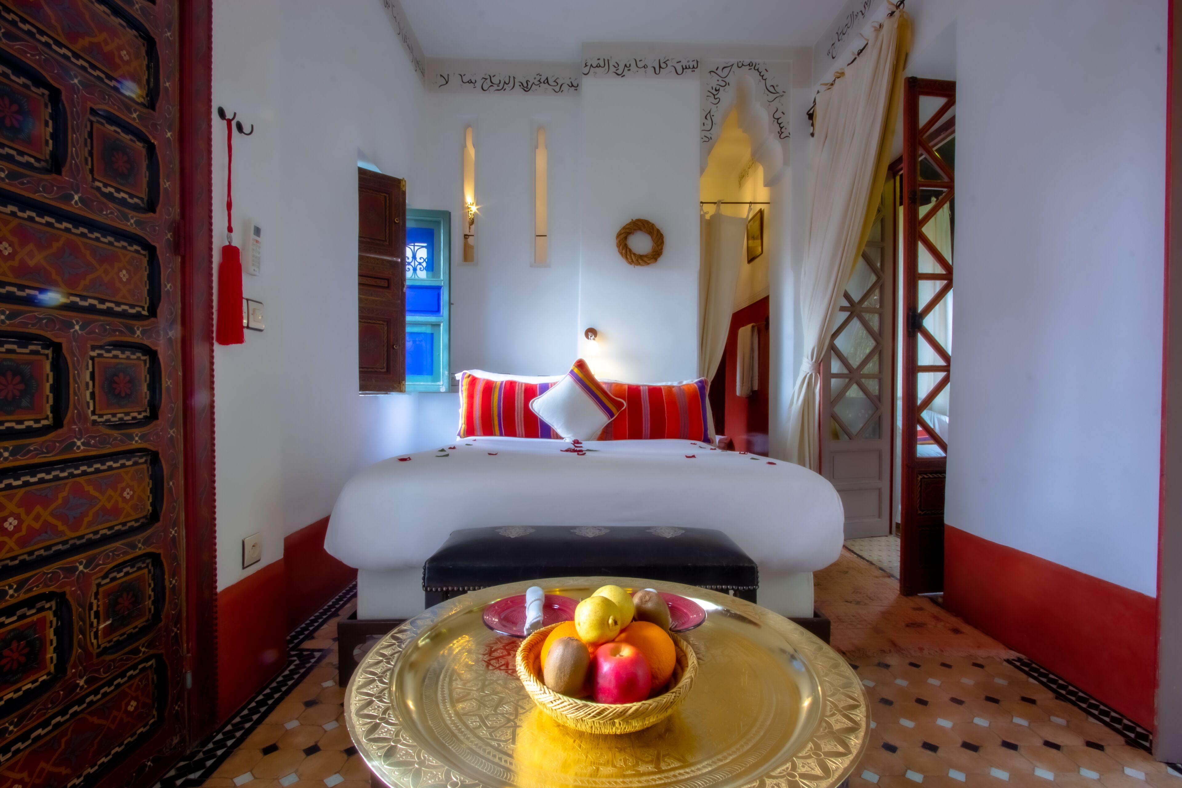 A hotel room with a bed, a fruit bowl on a tray, a red and white patterned door, and a blue window.