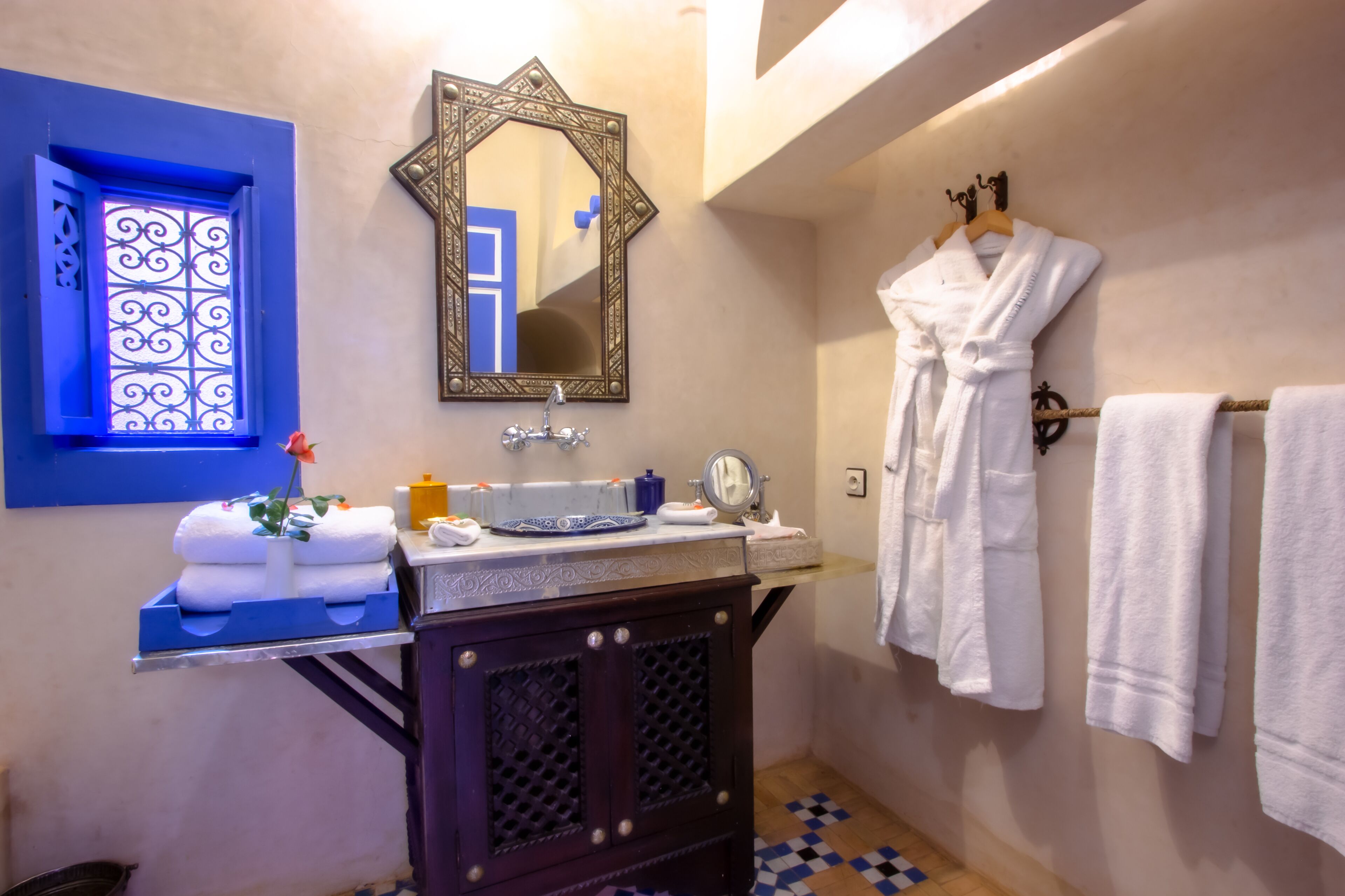 A bathroom with a blue window, a decorative mirror, a sink with toiletries, and a rack of white robes.