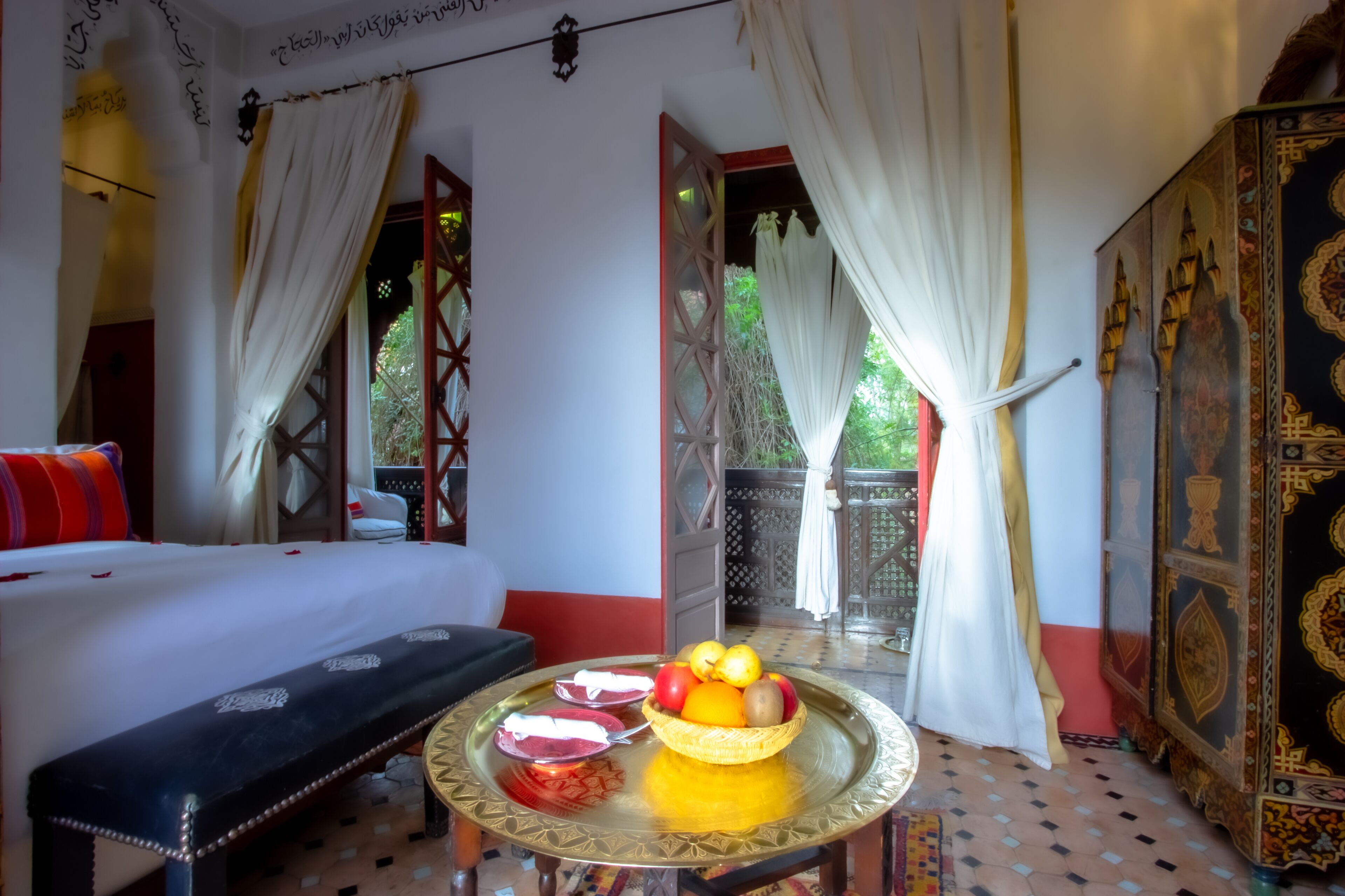 A room with a bed, a bench, a table with fruit, and a decorative cabinet.