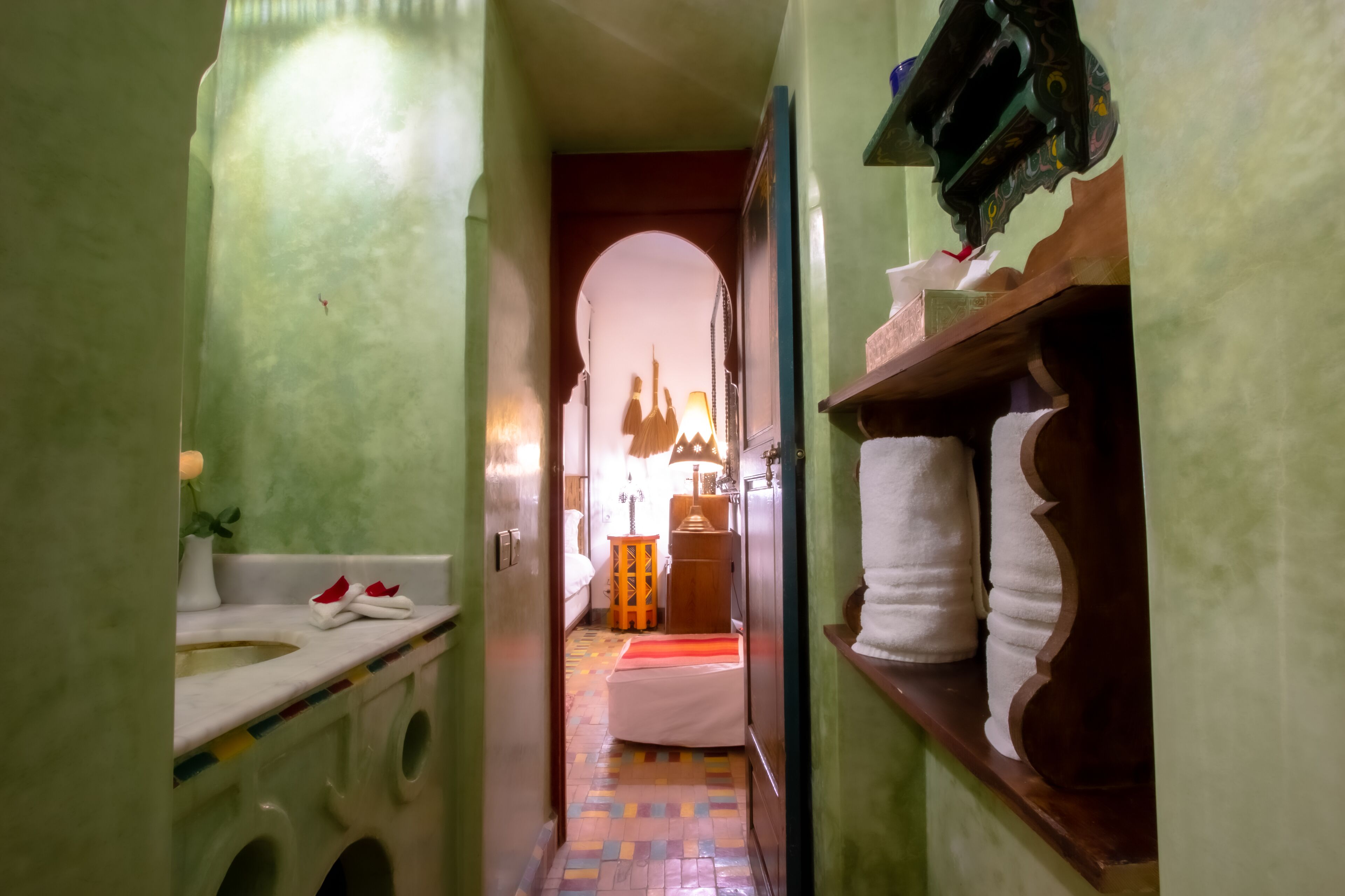 A narrow hallway with a bed in the distance, a sink on the left, and a shelf with towels on the right.