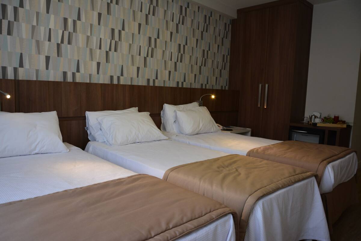 Quarto de hotel com três camas, cabeceira de madeira e parede estampada.