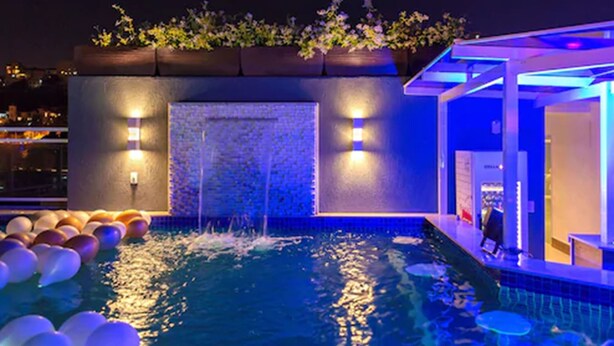 Une piscine sur le toit, agrémentée d’un élément décoratif aquatique, entourée de plantes en pot et éclairée par un système d’éclairage d’ambiance.