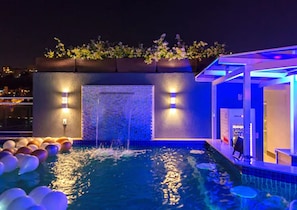 Ein Pool auf dem Dach mit Wasserspiel, umgeben von Blumentöpfen und beleuchtet durch stimmungsvolle Beleuchtung.