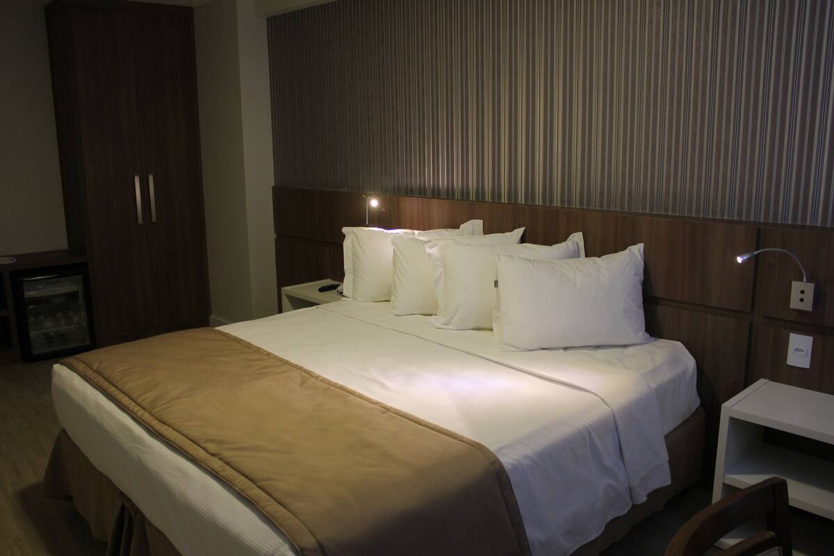 Quarto de hotel com uma cama grande, um guarda-roupa de madeira, uma mesa de cabeceira e um criado-mudo.