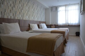 Ein Hotelzimmer mit zwei Betten, einem hölzernen Kopfteil, einer Wand mit Muster, einem Fenster mit durchscheinenden Vorhängen und einem kleinen Tisch mit einem Stuhl.