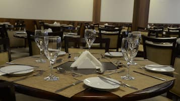 Una mesa de comedor dispuesta con copas de vino, platos, cubiertos y un portavasos para servilletas en un restaurante con varias mesas y sillas.