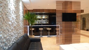 Un intérieur moderne avec un canapé en cuir noir, un coin bar doté de tabourets et un grand téléviseur à écran plat fixé sur un panneau en bois.