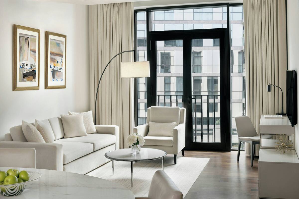 A modern living room with a sofa, armchair, coffee table, and a desk with a lamp. There are two framed artworks on the wall and a window with curtains.
