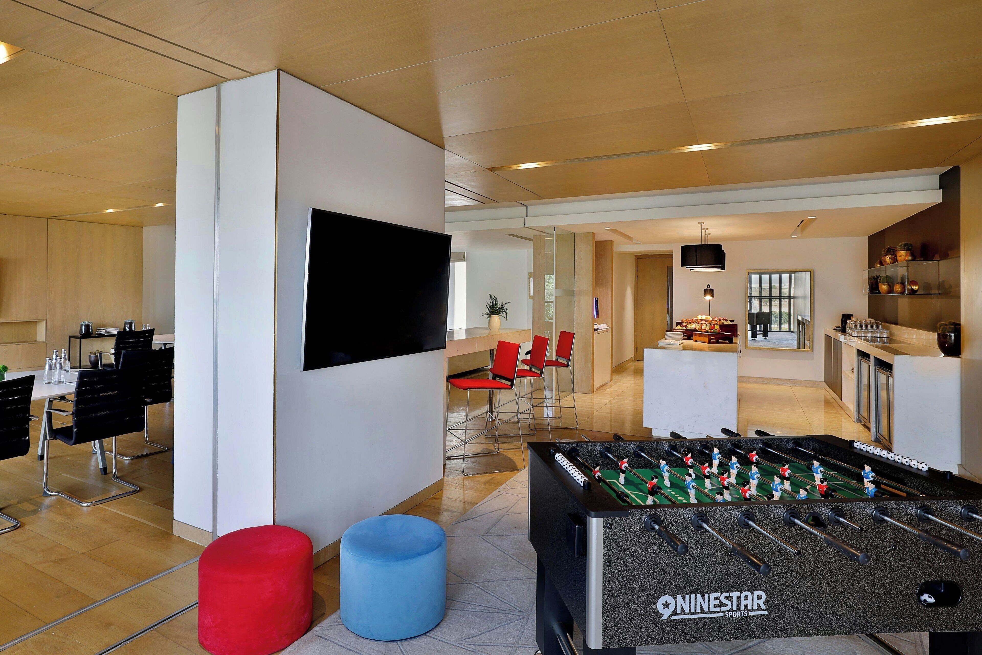 A modern hotel room with a foosball table, a flat-screen TV, and a bar area in the background.