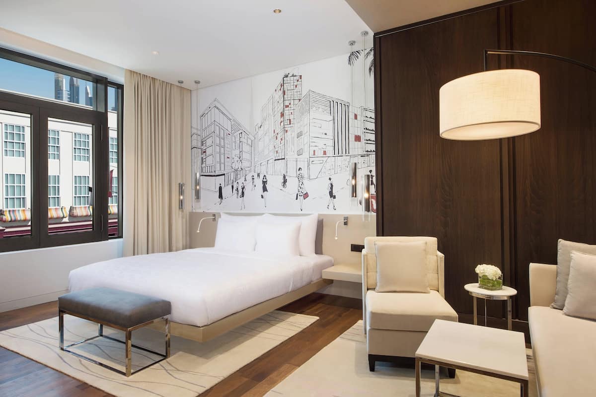 A modern hotel room with a large bed, a sofa, a small table, and a view of the cityscape through the window.