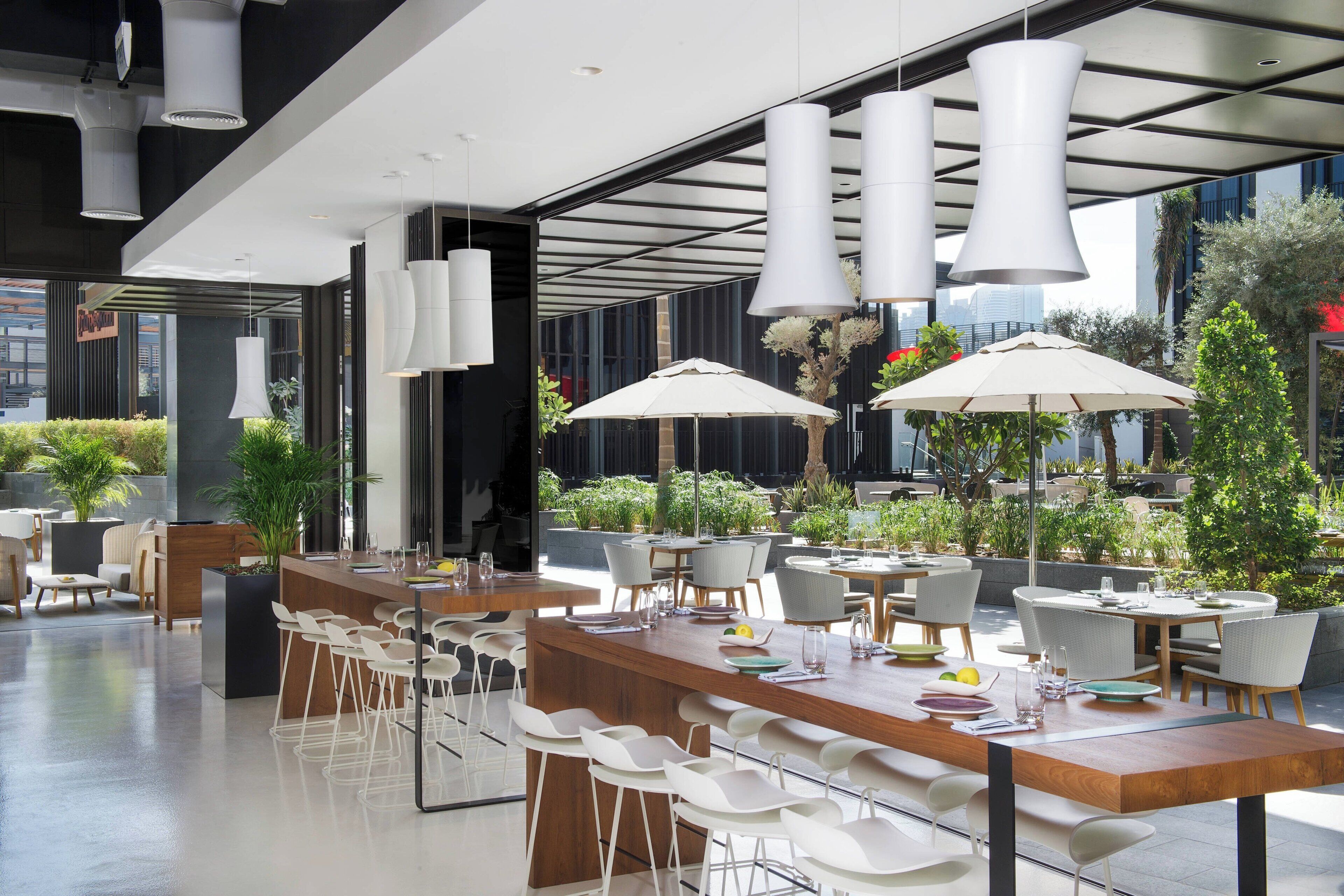 Un área de comedor al aire libre y moderna con sillas y mesas blancas, sombrillas grandes y plantas en macetas.