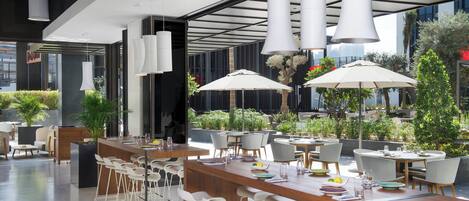 Un área de comedor al aire libre y moderna con sillas y mesas blancas, sombrillas grandes y plantas en macetas.
