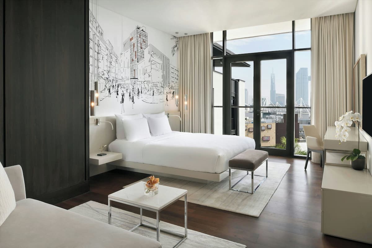 A modern hotel room with a large bed, a sitting area, a desk, and a view of the cityscape through large windows.
