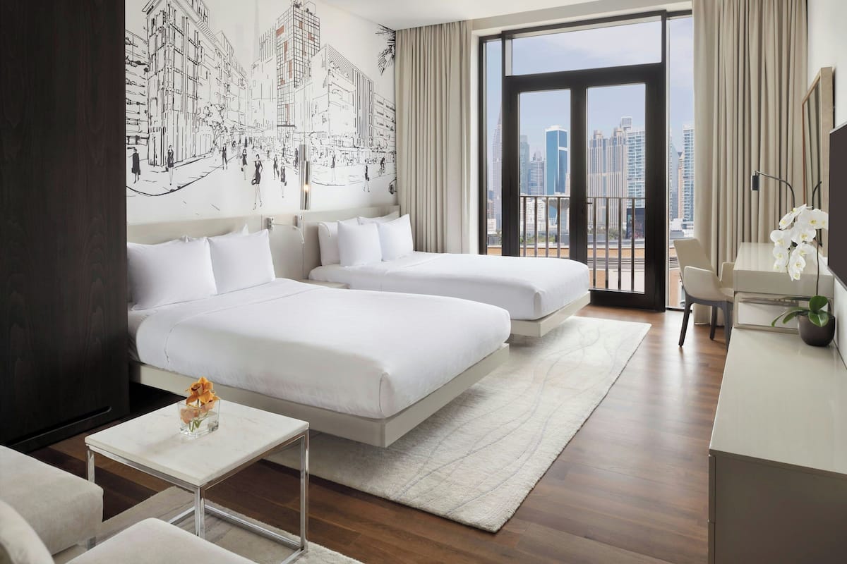 A hotel room with a large bed, a smaller bed, a desk, a chair, a TV, and a view of the city.