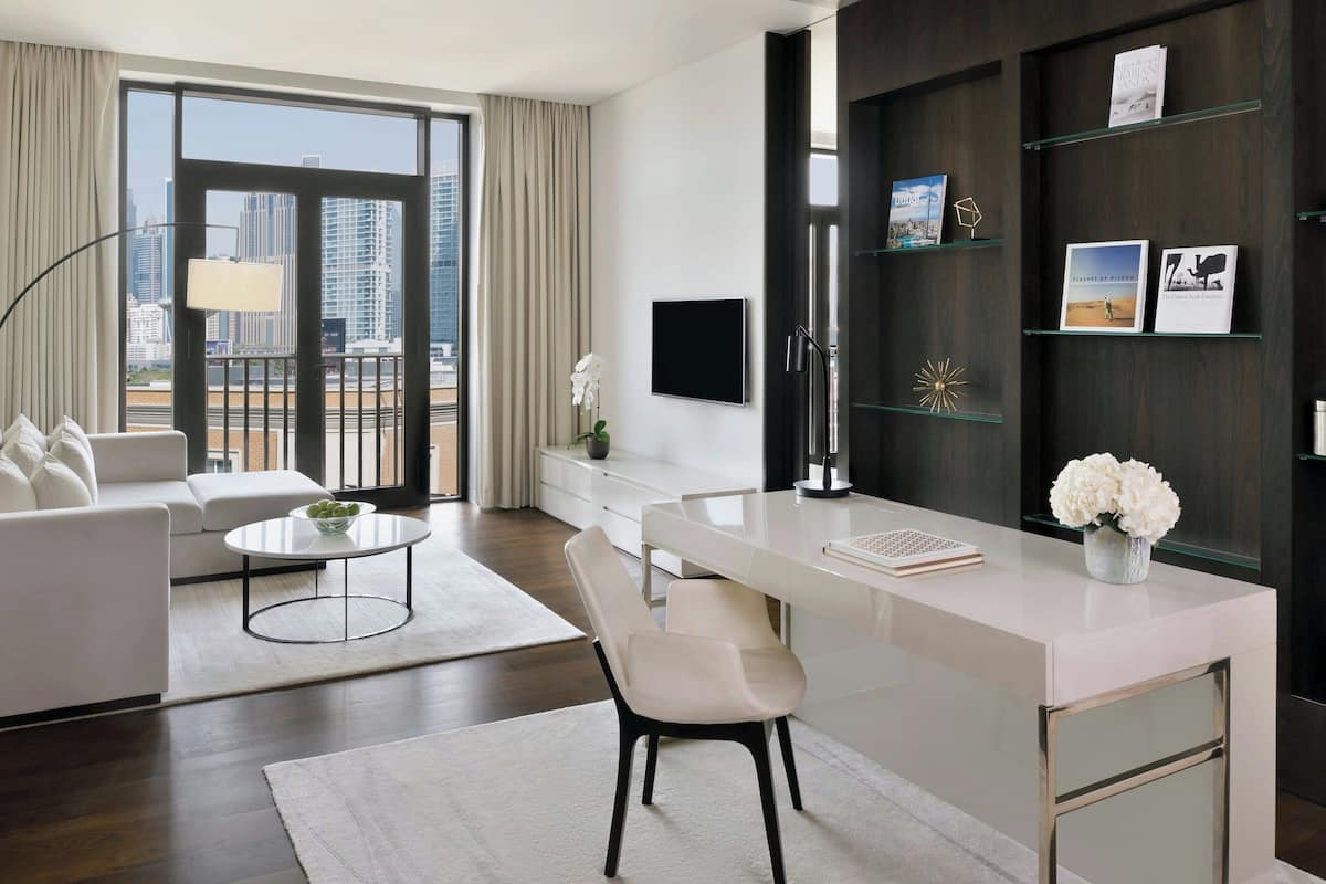 A modern hotel room with a large window, a desk with a chair, a sofa, and a bookshelf.