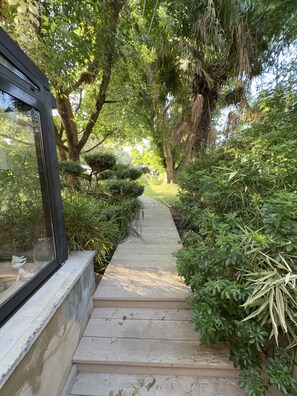 A wooden pathway flanked by dense greenery and a building with a glass window. - Le Corrigot (Pierry)