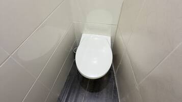 Un baño pequeño con azulejos blancos, un inodoro, un accionador de descarga en la pared y un accesorio para bidet metálico en el suelo.