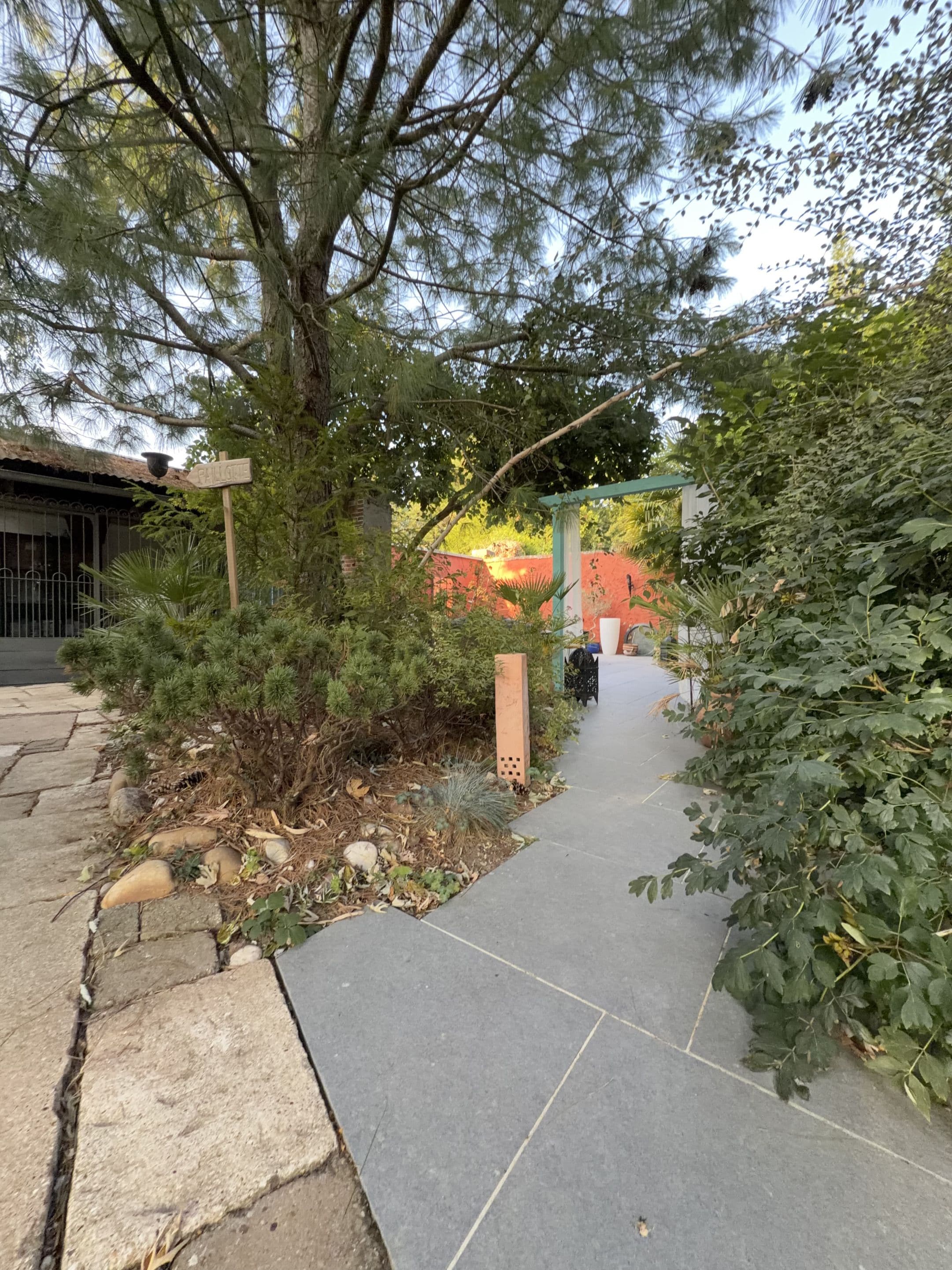 Un chemin menant à un bâtiment, agrémenté d’un panneau indicateur, et bordé de verdure et d’un mur rouge.