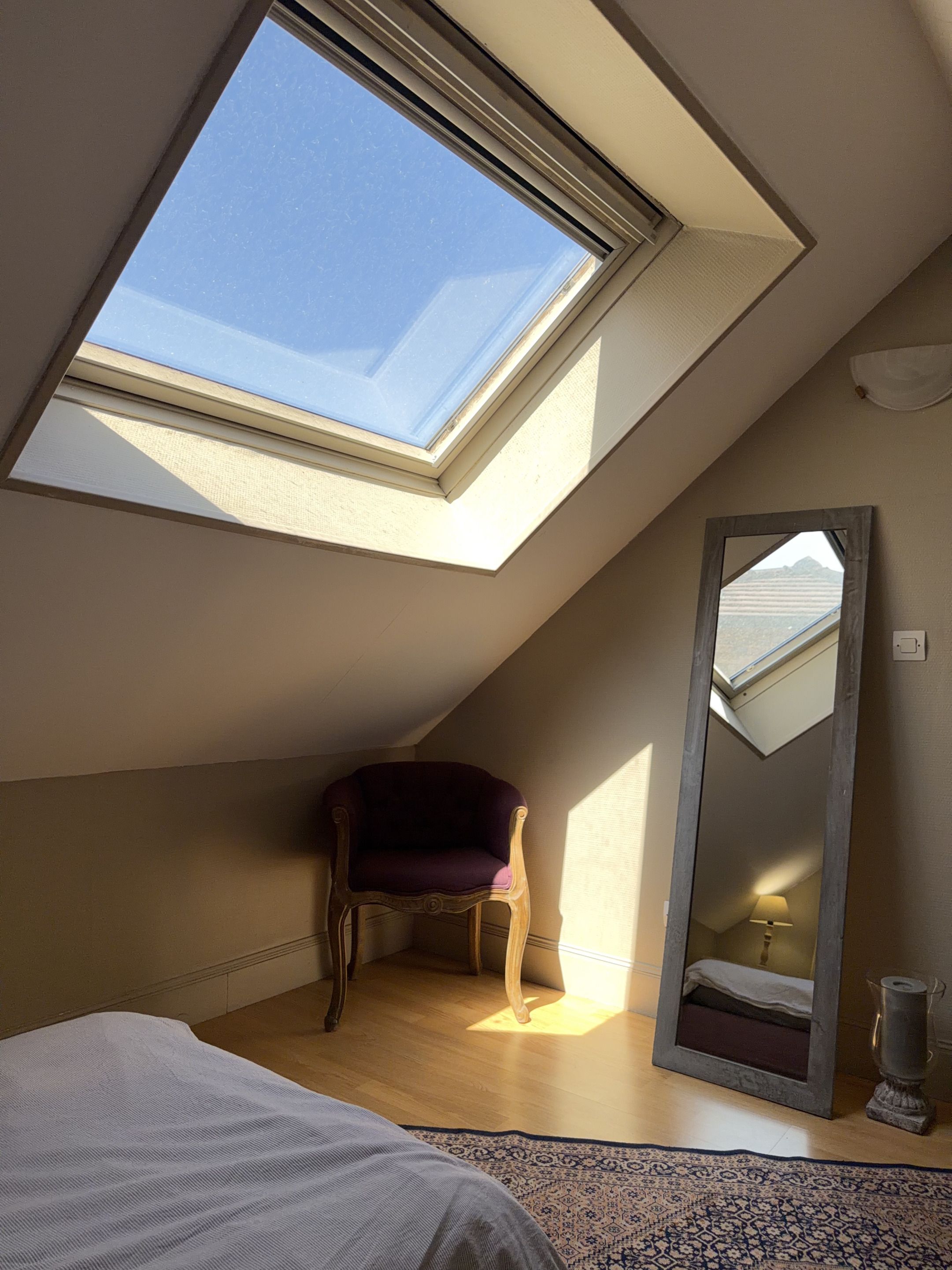 Una habitación con tragaluz, cama, silla, espejo y lámpara.