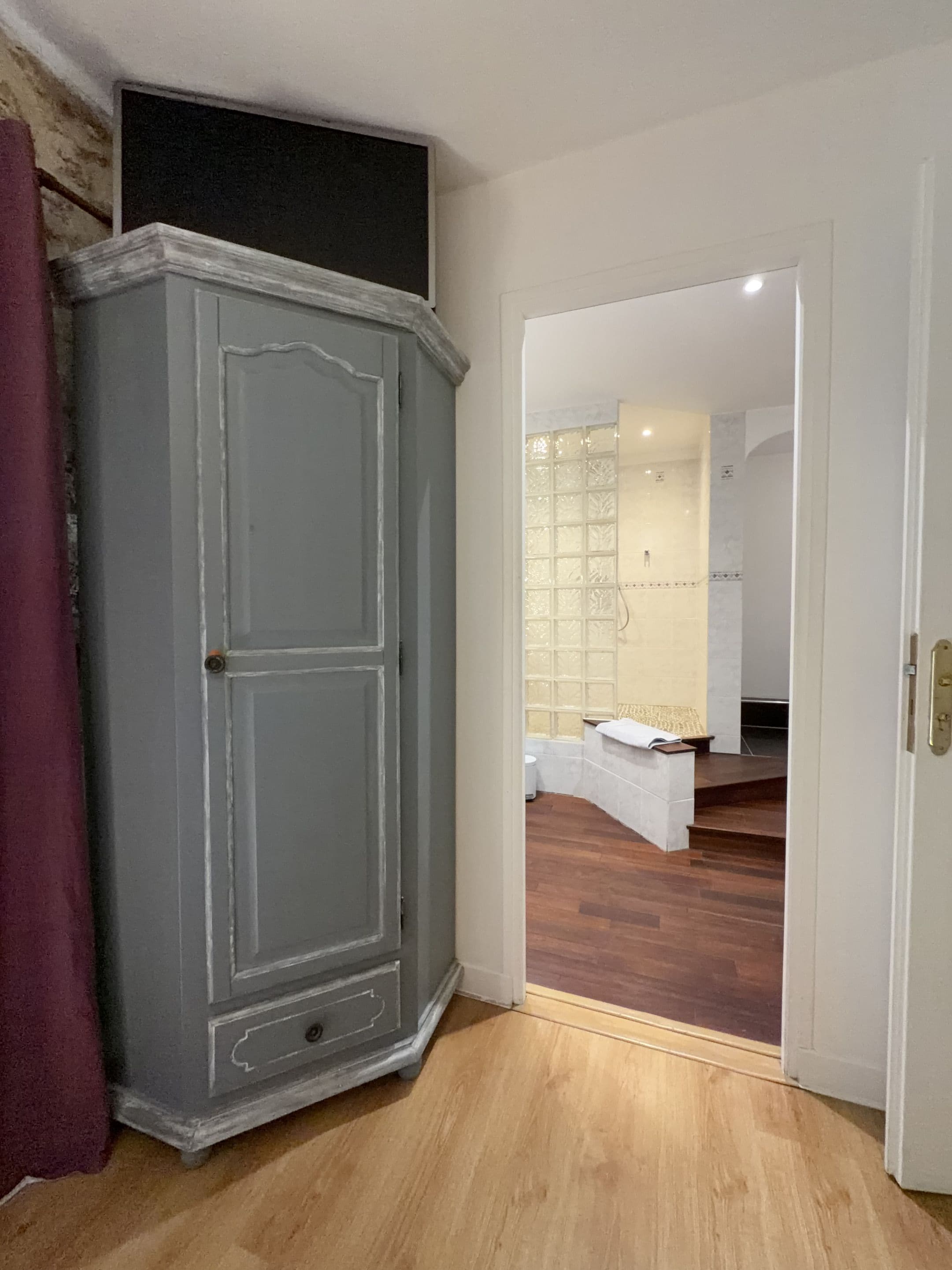A room with a grey wardrobe, a white door leading to a bathroom with a glass shower enclosure.