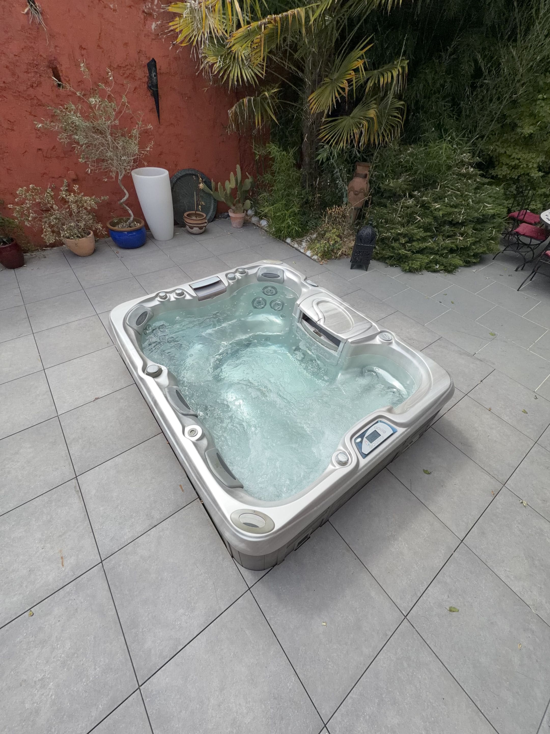Een bubbelbad in een openlucht setting met betegelde vloer, omringd door potplanten en een rode muur.