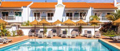 A swimming pool with lounge chairs and umbrellas, a white building with orange tiled roofs, and a clear blue sky.