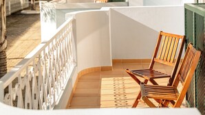 A balcony with white railings, two wooden chairs, and a view of a garden with palm trees and a building in the background. - Giramar Apartamentos Turisticos (Lagos)