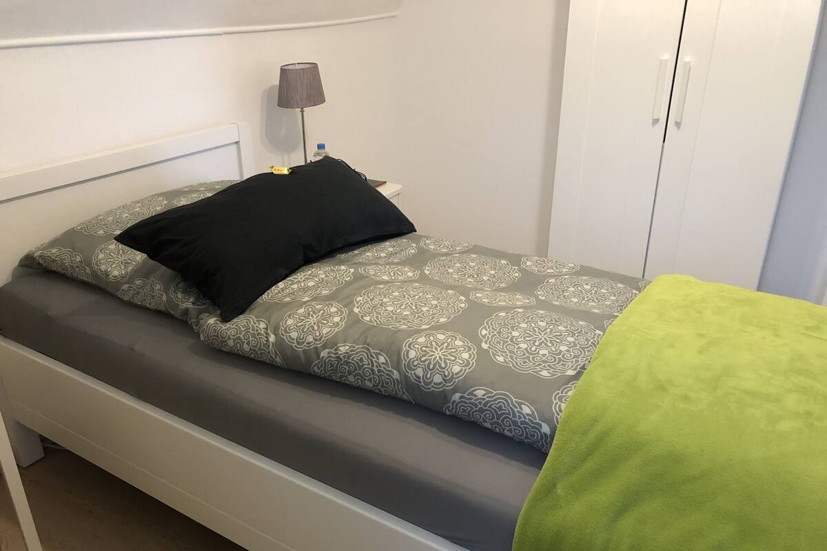 A single bed with a grey patterned bedspread and a black pillow, a grey lamp, and a white wardrobe in a room with white walls.