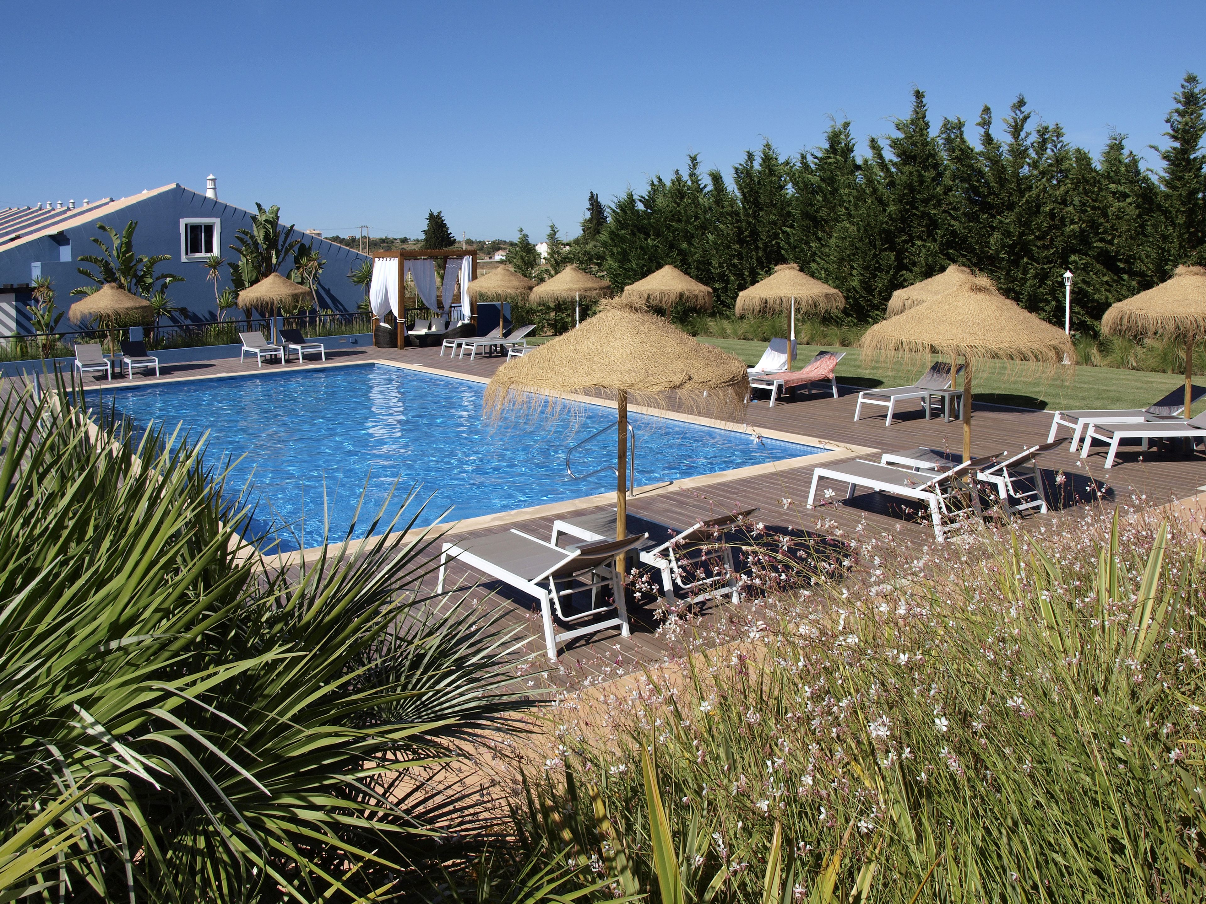 A swimming pool with lounge chairs and thatched umbrellas, surrounded by greenery and a building in the background.