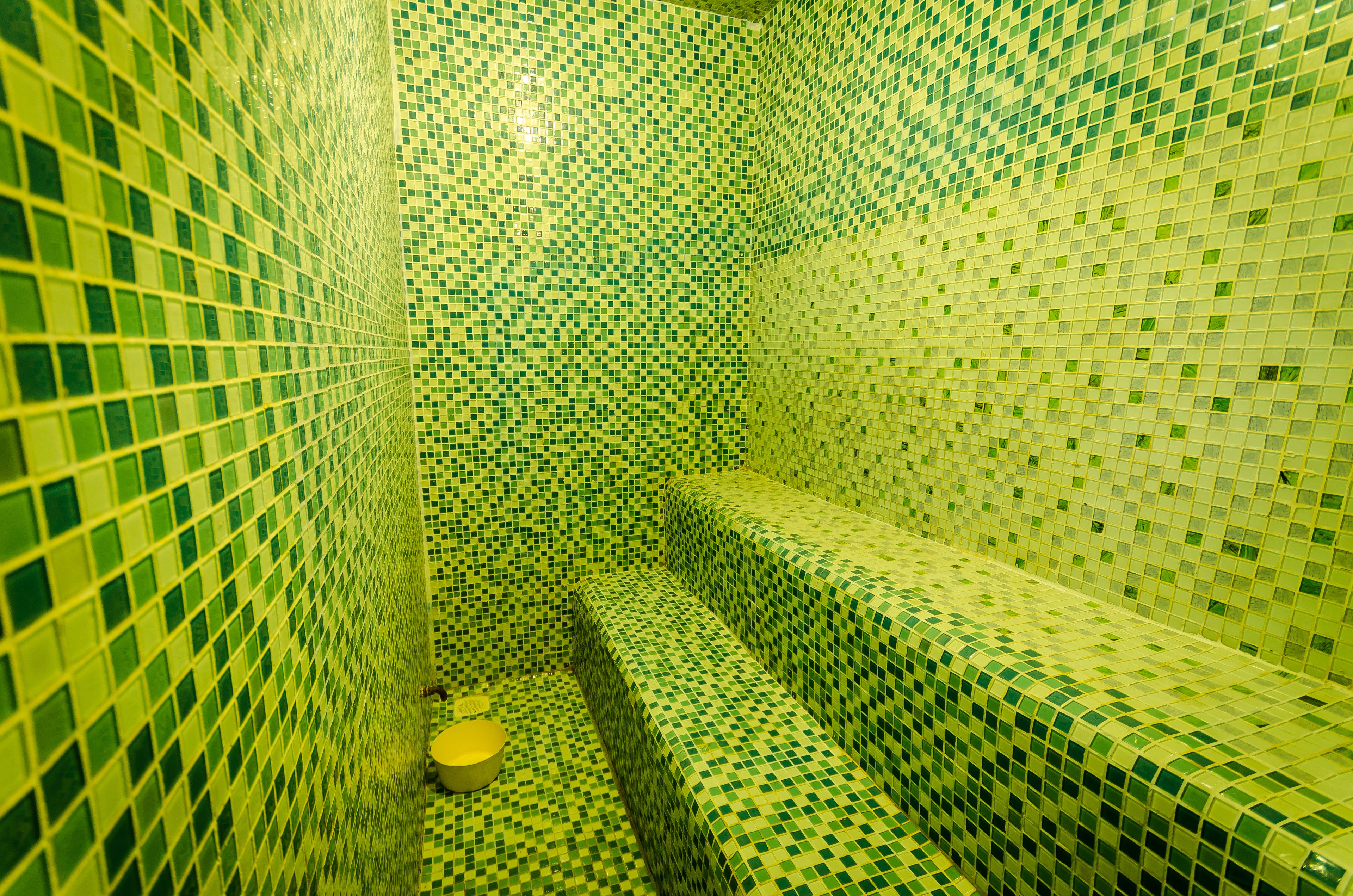 Une salle de bain avec des murs et un sol recouverts de mosaïques vertes et blanches, un lavabo jaune et un banc.
