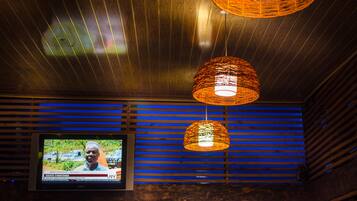 Una habitación con televisión mostrando un noticiero, techo de listones de madera y dos lámparas colgantes.